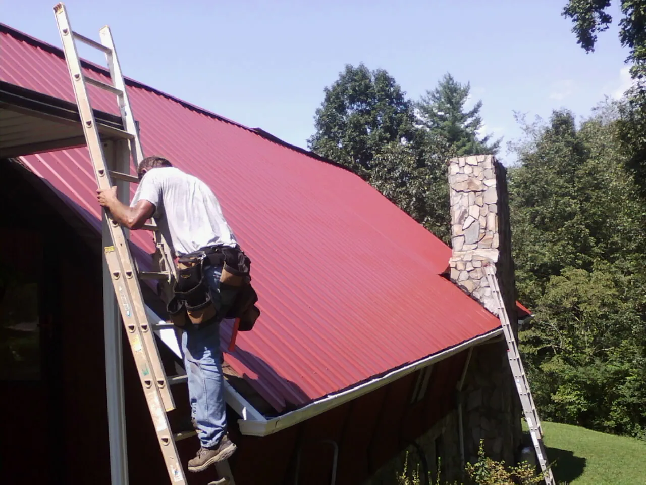 Roofer on a ladder beside a red metal roof and stone chimney — Soffit & Fascia Repair by Roofing Brothers in Lewellen
